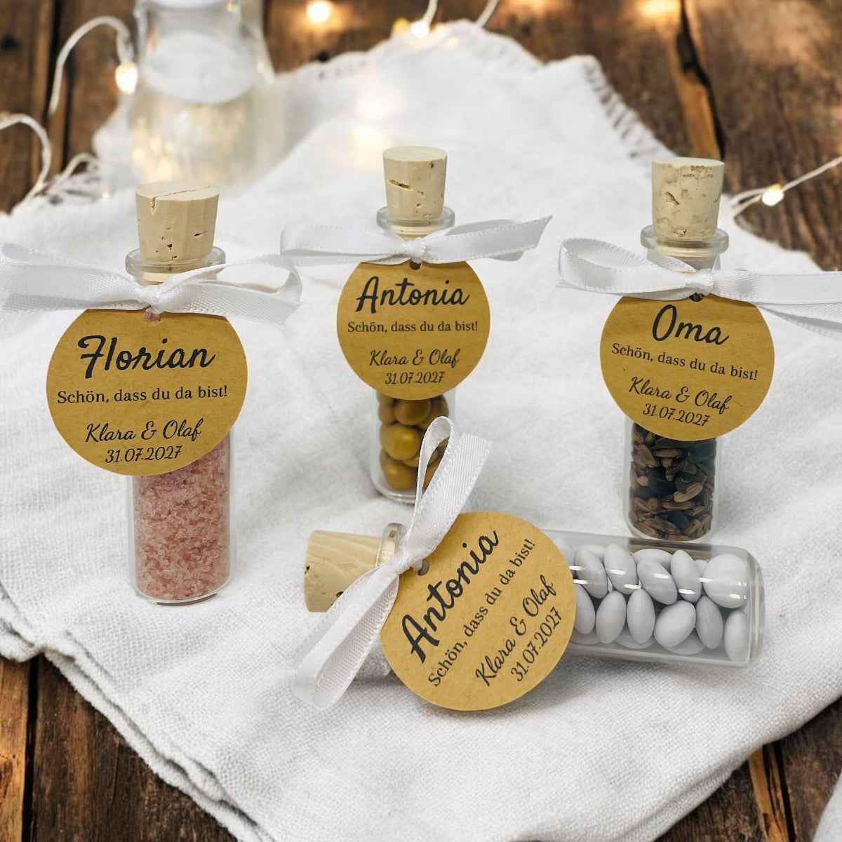 Vier personalisierte Gastgeschenke aus kleinen Glas-Schiffchen mit Naturkorken stehen auf einer festlich gedeckten weißen Tafel. An jedem Glas ist mit einem weißen Satinband eine runde Kraftpapier-Tischkarte befestigt. Die Gläser sind mit rosa Badesalz, Schokolinsen, Sonnenblumenkernen und weißen Dragees gefüllt und dienen gleichzeitig als elegante Tischkarten für Hochzeit, Taufe oder Geburtstag.