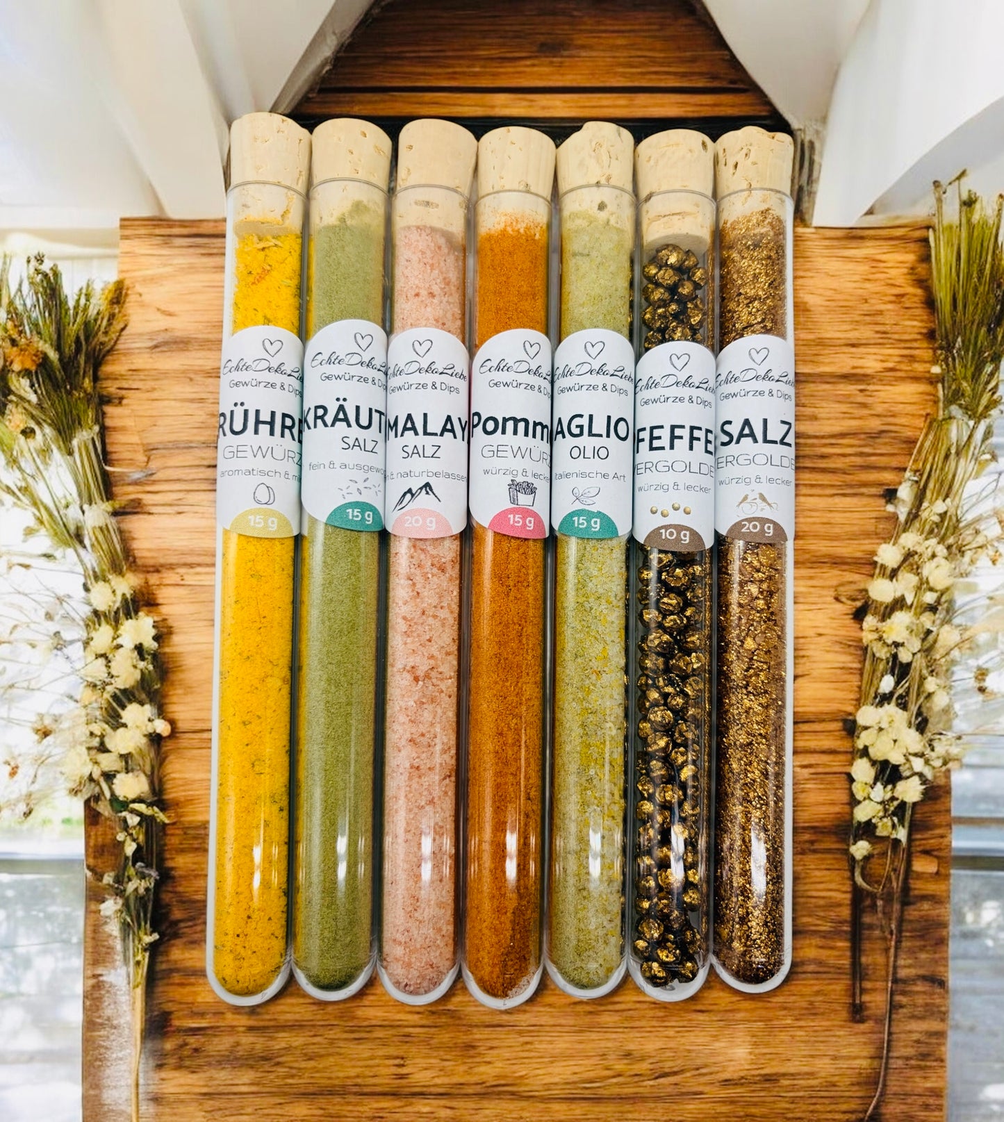 Sieben Reagenzgläser mit Naturkorken, gefüllt mit verschiedenen Gewürzen und Salzen. Zu sehen sind Rühreigewürz, Kräutersalz, Himalayasalz, Pommesgewürz, Aglio-Olio-Gewürz, vergoldeter Pfeffer und vergoldetes Salz. Die Gläser tragen moderne weiße Etiketten mit Produktname und Füllmenge. Arrangiert auf hellem Holzhintergrund mit dekorativen Trockenblumen – eine besondere Geschenkidee für Gewürzliebhaber.