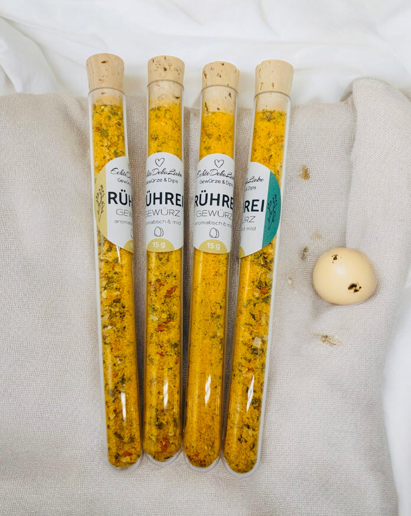 Sieben Reagenzgläser mit Naturkorken, gefüllt mit verschiedenen Gewürzen und Salzen. Zu sehen sind Rühreigewürz, Kräutersalz, Himalayasalz, Pommesgewürz, Aglio-Olio-Gewürz, vergoldeter Pfeffer und vergoldetes Salz. Die Gläser tragen moderne weiße Etiketten mit Produktname und Füllmenge. Arrangiert auf hellem Holzhintergrund mit dekorativen Trockenblumen – eine besondere Geschenkidee für Gewürzliebhaber.