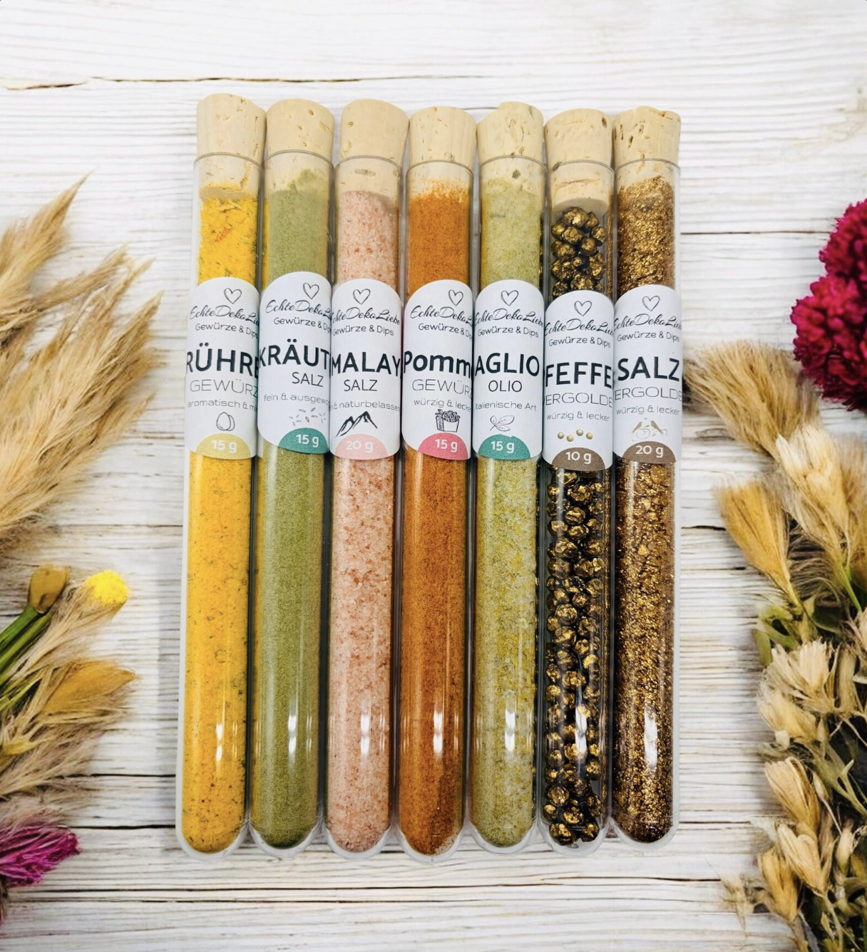 Sieben Reagenzgläser mit Naturkorken, gefüllt mit verschiedenen Gewürzen und Salzen. Zu sehen sind Rühreigewürz, Kräutersalz, Himalayasalz, Pommesgewürz, Aglio-Olio-Gewürz, vergoldeter Pfeffer und vergoldetes Salz. Die Gläser tragen moderne weiße Etiketten mit Produktname und Füllmenge. Arrangiert auf hellem Holzhintergrund mit dekorativen Trockenblumen – eine besondere Geschenkidee für Gewürzliebhaber.