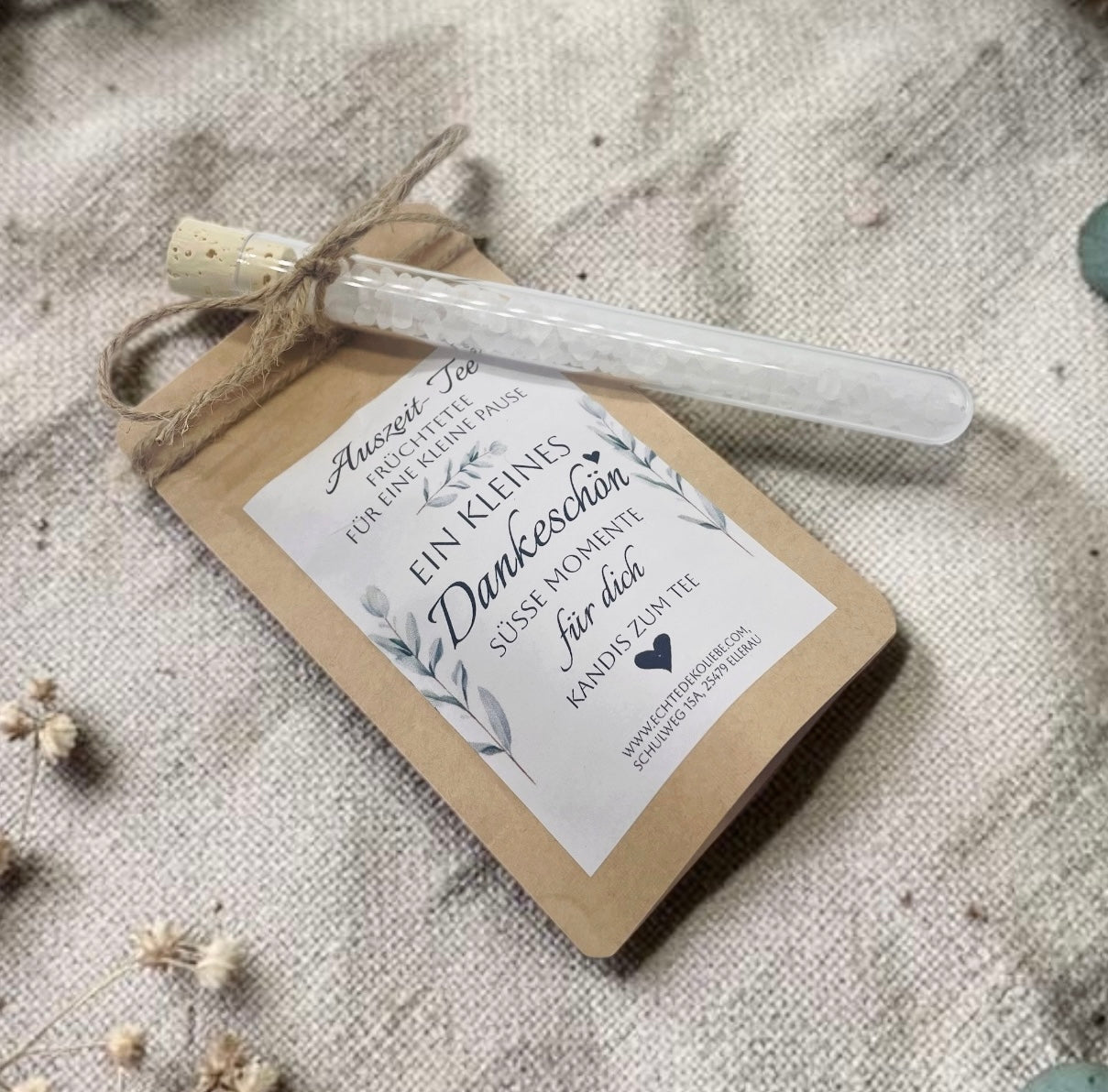 Braune Kraftpapiertüte mit aromatischem Früchtetee und einem weißen Etikett im zarten Eukalyptus-Design. Aufdruck: „Auszeit-Tee – Früchtetee für eine kleine Pause. Ein kleines Dankeschön. Süße Momente für dich. Kandis zum Tee.“ Mit Bastband ist ein Reagenzglas aus Glas befestigt, das mit weißem Kandis gefüllt ist. Das Geschenk liegt dekorativ auf einer rustikalen Holzoberfläche neben einem hellen Leinentuch und vermittelt eine natürliche, liebevolle Geschenkidee.