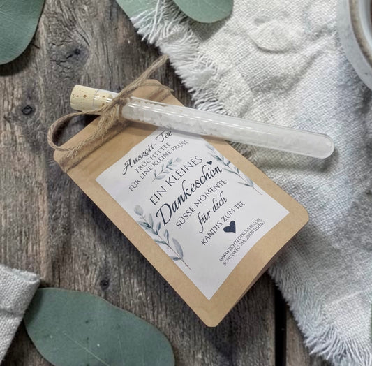 Braune Kraftpapiertüte mit aromatischem Früchtetee und einem weißen Etikett im zarten Eukalyptus-Design. Aufdruck: „Auszeit-Tee – Früchtetee für eine kleine Pause. Ein kleines Dankeschön. Süße Momente für dich. Kandis zum Tee.“ Mit Bastband ist ein Reagenzglas aus Glas befestigt, das mit weißem Kandis gefüllt ist. Das Geschenk liegt dekorativ auf einer rustikalen Holzoberfläche neben einem hellen Leinentuch und vermittelt eine natürliche, liebevolle Geschenkidee.