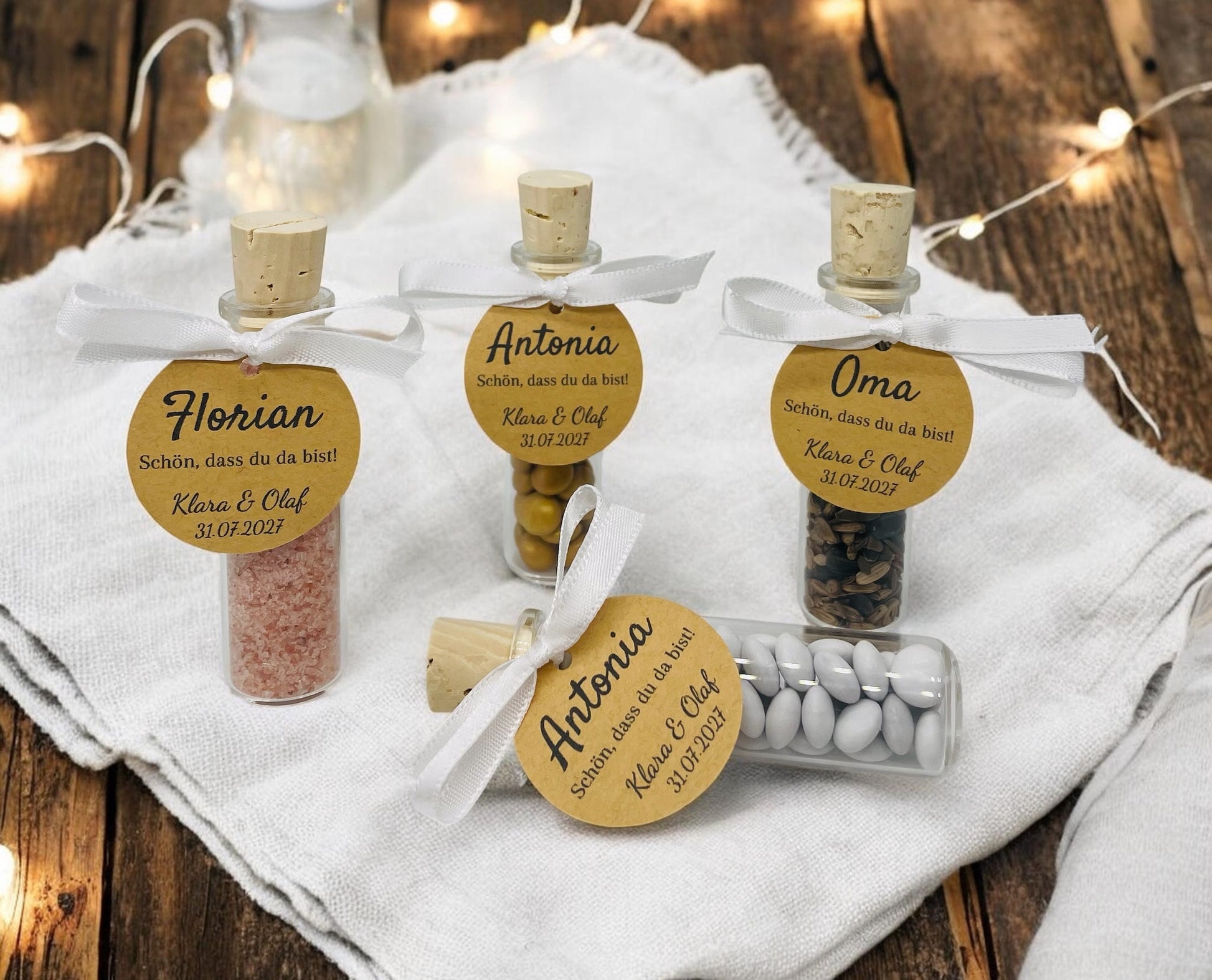 Vier personalisierte Gastgeschenke aus kleinen Glas-Schiffchen mit Naturkorken stehen auf einer festlich gedeckten weißen Tafel. An jedem Glas ist mit einem weißen Satinband eine runde Kraftpapier-Tischkarte befestigt. Die Gläser sind mit rosa Badesalz, Schokolinsen, Sonnenblumenkernen und weißen Dragees gefüllt und dienen gleichzeitig als elegante Tischkarten für Hochzeit, Taufe oder Geburtstag.