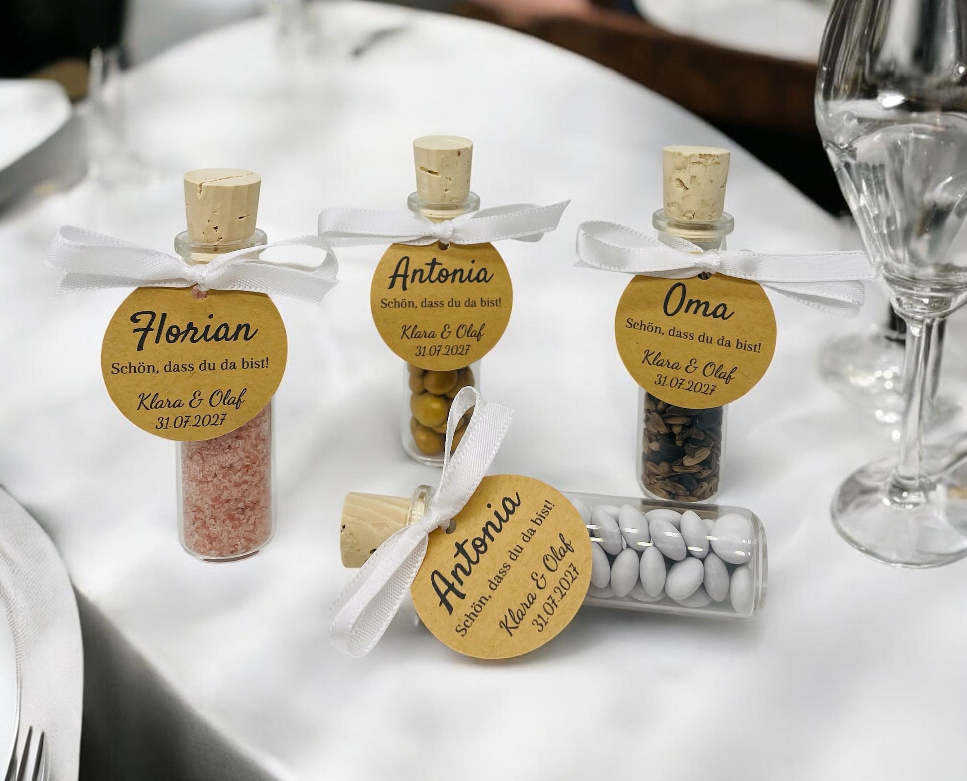 Vier personalisierte Gastgeschenke aus kleinen Glas-Schiffchen mit Naturkorken stehen auf einer festlich gedeckten weißen Tafel. An jedem Glas ist mit einem weißen Satinband eine runde Kraftpapier-Tischkarte befestigt. Die Gläser sind mit rosa Badesalz, Schokolinsen, Sonnenblumenkernen und weißen Dragees gefüllt und dienen gleichzeitig als elegante Tischkarten für Hochzeit, Taufe oder Geburtstag.
