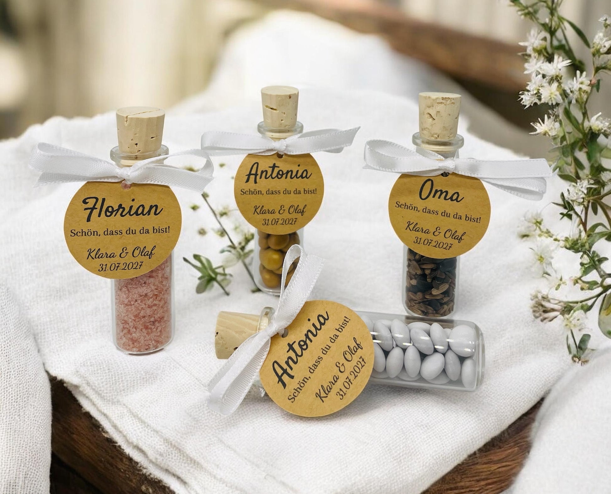 Vier personalisierte Gastgeschenke aus kleinen Glas-Schiffchen mit Naturkorken stehen auf einer festlich gedeckten weißen Tafel. An jedem Glas ist mit einem weißen Satinband eine runde Kraftpapier-Tischkarte befestigt. Die Gläser sind mit rosa Badesalz, Schokolinsen, Sonnenblumenkernen und weißen Dragees gefüllt und dienen gleichzeitig als elegante Tischkarten für Hochzeit, Taufe oder Geburtstag.