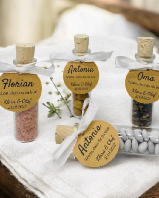 Vier personalisierte Gastgeschenke aus kleinen Glas-Schiffchen mit Naturkorken stehen auf einer festlich gedeckten weißen Tafel. An jedem Glas ist mit einem weißen Satinband eine runde Kraftpapier-Tischkarte befestigt. Die Gläser sind mit rosa Badesalz, Schokolinsen, Sonnenblumenkernen und weißen Dragees gefüllt und dienen gleichzeitig als elegante Tischkarten für Hochzeit, Taufe oder Geburtstag.