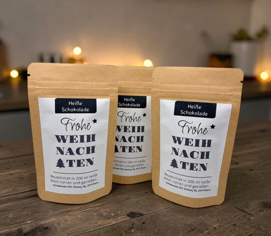 Braune Kraftpapiertüten mit weihnachtlichem Design stehen dekorativ auf einem rustikalen Holztisch. Die Tüten sind mit einem winterlich gestalteten Etikett versehen und enthalten eine Getränkemischung bzw. ein Heißgetränk zum Genießen. Im Hintergrund sorgen Tannenzweige, warme Lichter und weihnachtliche Dekoration für eine gemütliche Adventsstimmung.